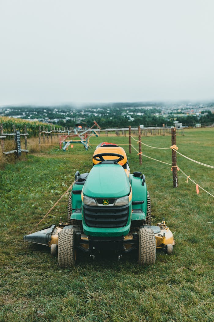 A green riding lawn mower sits on a vast grassy field with rustic fencing in a tranquil rural setting.